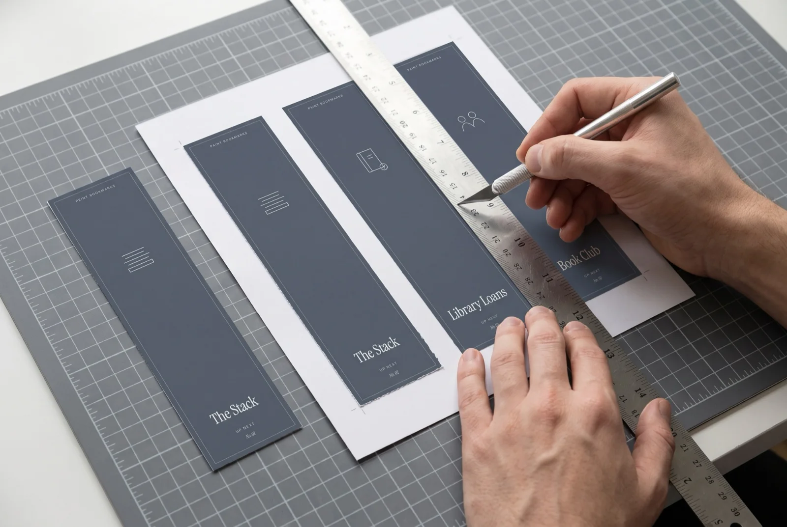 Hands trimming a printed sheet of bookmarks with a ruler and craft knife.