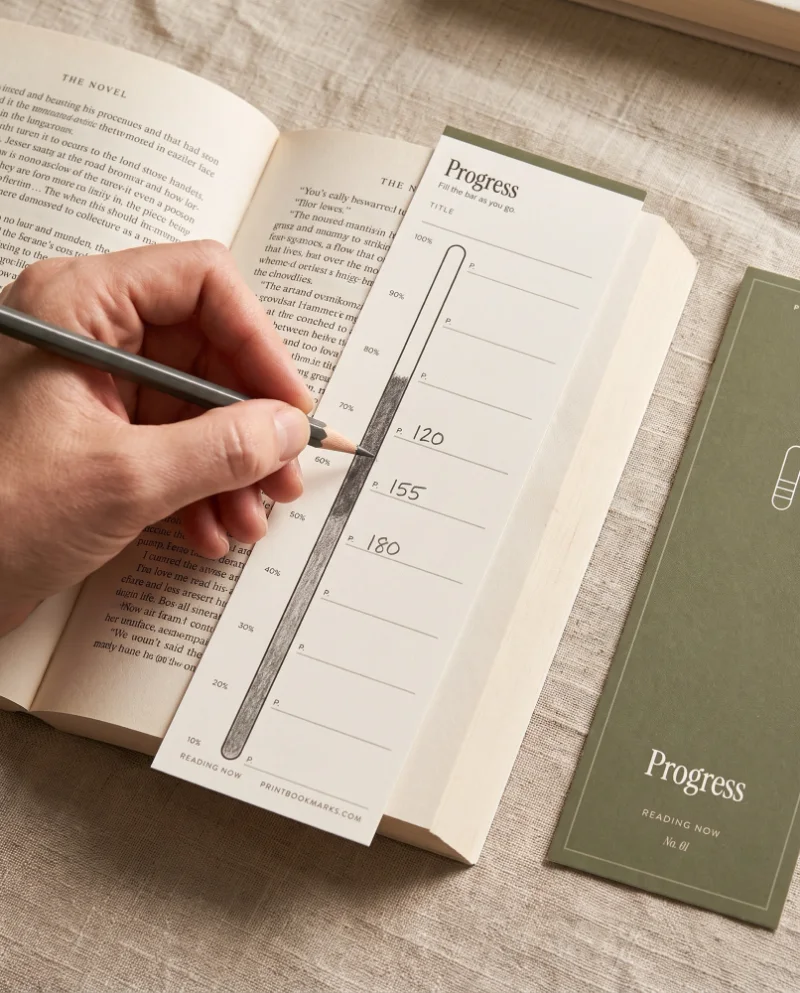A hand writing page numbers in pencil beside a shaded progress bar on a cream Progress bookmark lying across an open paperback, with the olive green front of the same bookmark on the linen beside it.