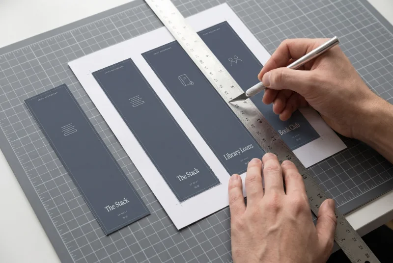 Hands cutting a printed sheet of dark blue bookmarks with a craft knife along a steel ruler on a gray cutting mat, with two cut bookmarks titled The Stack beside it